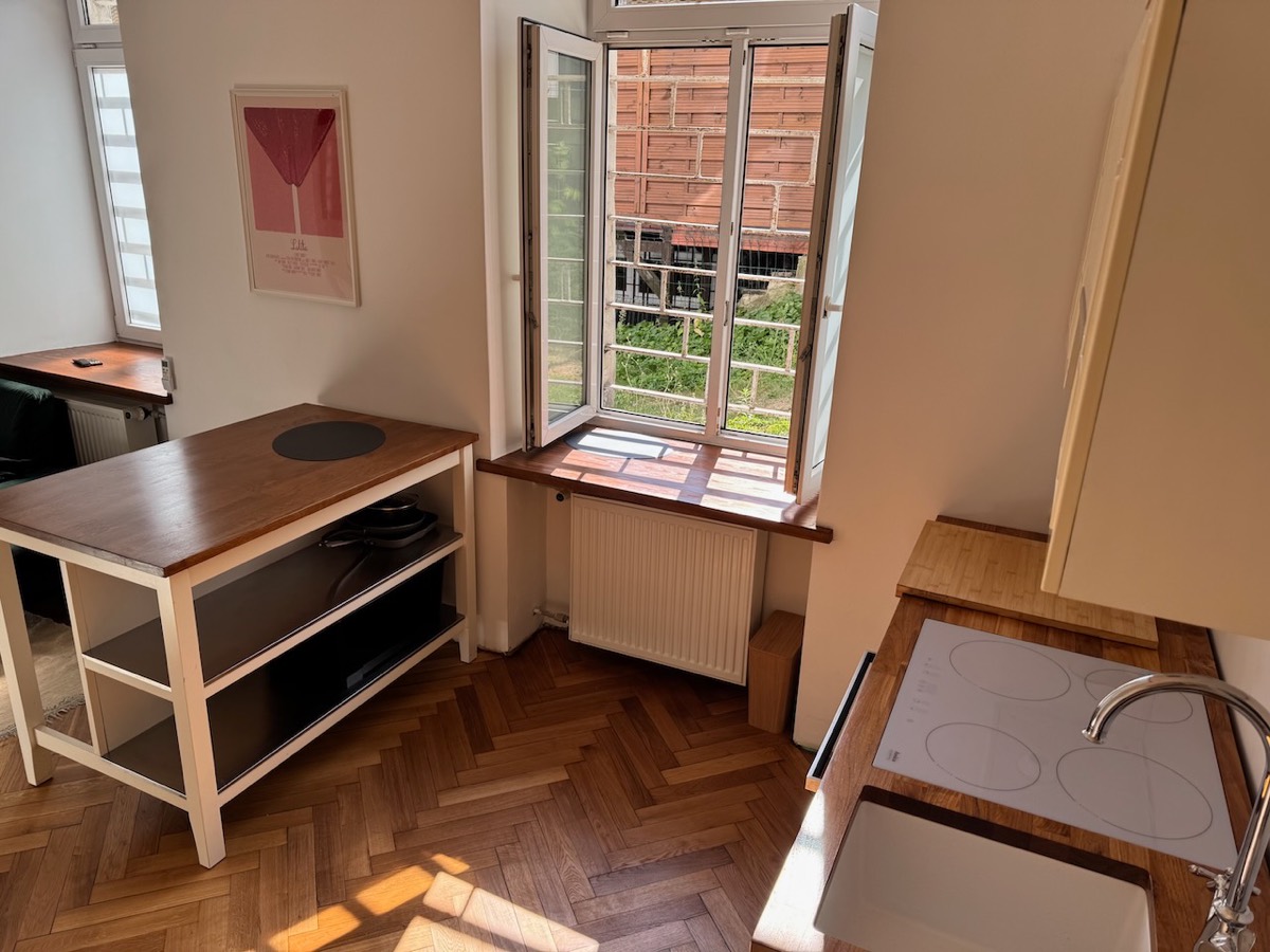 Sunlit kitchen with a tall casement window open onto courtyard greenery and oak herringbone floor